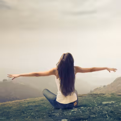 A Woman is Sitting on Grass in the Mountains with Arms Stretched Out and Looking at the Grey Sky