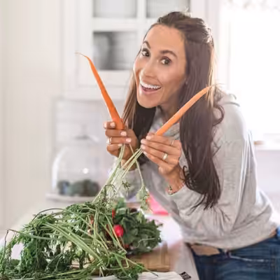 Female Wearing Grey Hoodie in Kitchen Smiling and Posing While Holding Two Carrots