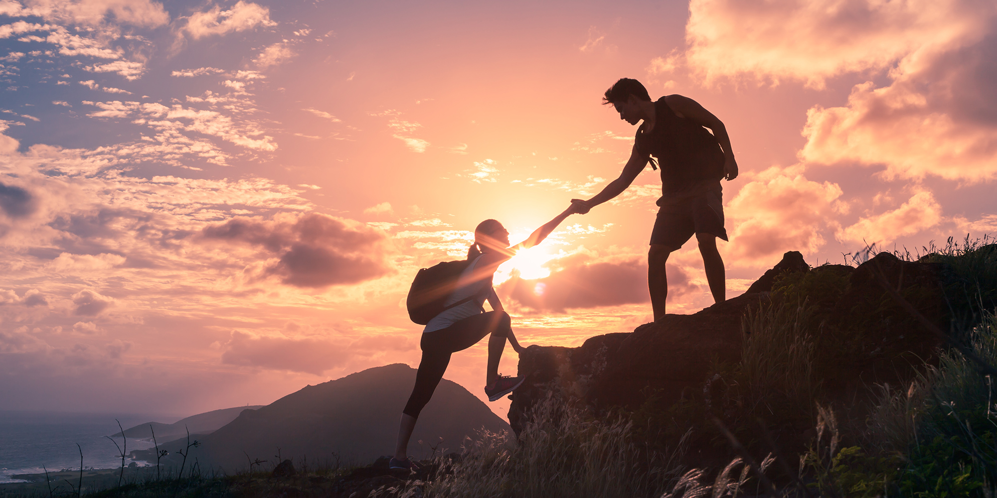 Man is Helping Woman to Climb on a Cliff In Front of a Beautiful Sky and Sun Rays
