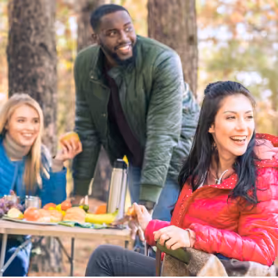 Group of People Sitting Around the Table in Nature and Eating Fruits While Looking in the Distance and Smiling