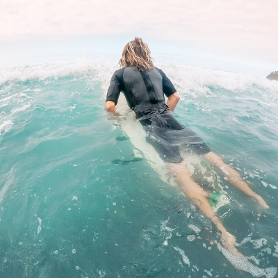 Woman in Black Surf Clothing is Laying on the Surfboard in the Sea