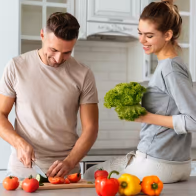 A Happy Man is Slicing Vegetables in the Kitchen and a Woman is Sitting on the Kitchen Countertop Smiling and Holding Broccoli