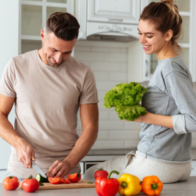 A Happy Man is Slicing Vegetables in the Kitchen and a Woman is Sitting on the Kitchen Countertop Smiling and Holding Broccoli