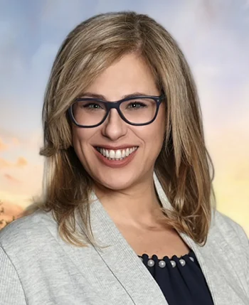 A professional headshot of a smiling woman with blonde hair wearing glasses, a grey blazer, and a navy top, set against a sunset sky background.