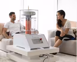 A man and a woman seated in a modern living room, smiling and engaging in a lively conversation with a cutting-edge home device placed on a coffee table between them.