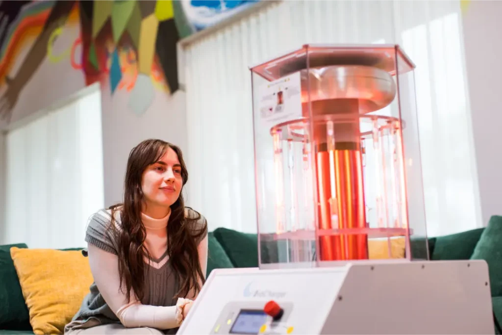 young, brunette woman staring warmly at a health optimization device called the BioCharger in front of her in a well lit area on a green couch with yellow pillows