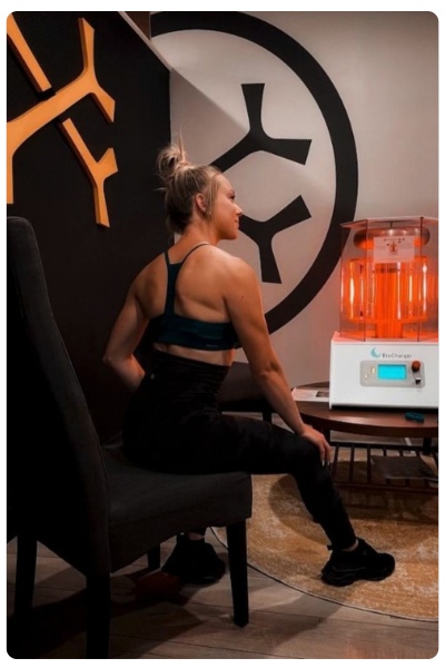 Fit women in a black workout outfit sitting in front of a brightly lit BioCharger and OsteoStrong graphics