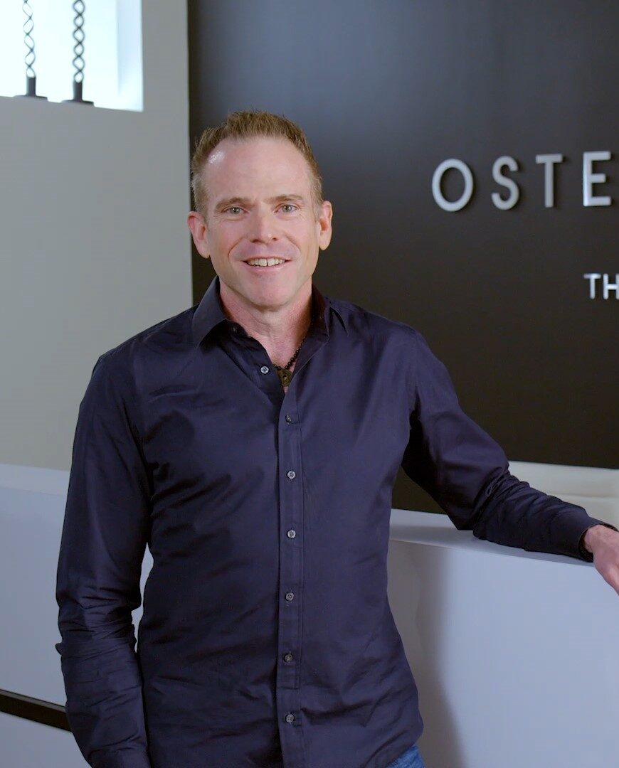 Man in a blue button-down shirt standing at a white desk in front of a white OsteoStrong sign